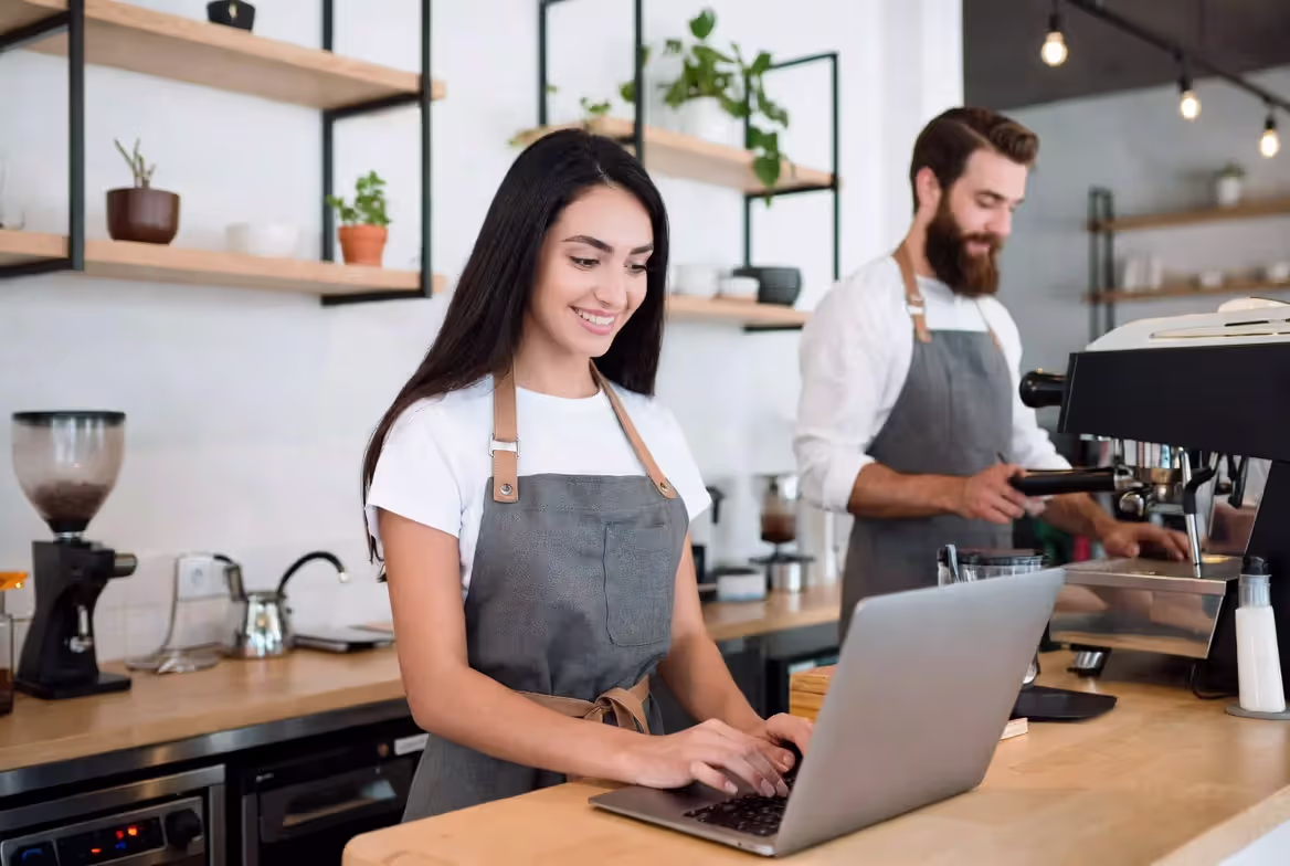 Smiling young woman with long dark hair working on a laptop at a modern cafe counter, with a male barista preparing coffee in the background